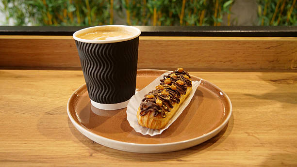 istock Creamy foam on cappuccino cup with french chocolate eclair topped with nuts on a table in confectionery store 2233980155