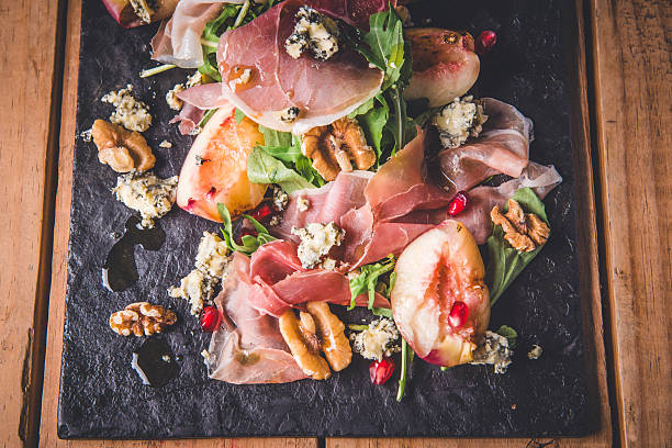 A delicious and fresh gourmet salad featuring thinly sliced prosciutto, juicy peach slices, and crumbled blue cheese. The salad is enhanced with arugula, walnuts, and pomegranate seeds, creating a vibrant presentation. Served on a dark slate plate, it embodies elegance and sophistication, perfect for a gourmet menu or food presentation.