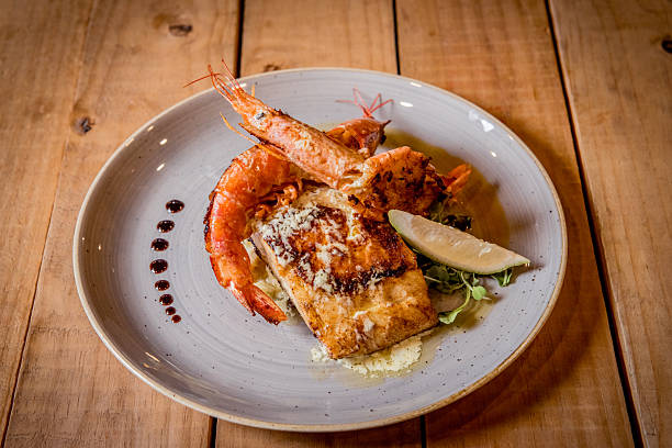 Gourmet seafood cuisine featuring grilled shrimp and fish fillets, arranged artistically on a plate with garnishes including a lemon wedge and a decorative sauce drizzle. This appetizing dish is served on a rustic wooden table, enhancing the warm and inviting aesthetics of the presentation.