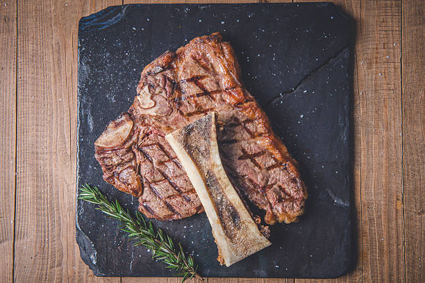 A delectable grilled steak served on a black slate platter with a rosemary garnish on a rustic wooden surface. The image showcases grill marks and a bone piece, emphasizing the art of cooking. Perfect for culinary and food-themed concepts.