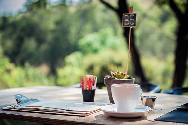 Elegant outdoor cafe table setup featuring a white coffee cup, succulent plant, and menus, surrounded by lush greenery. The scene exudes comfort and relaxation, perfect for enjoying a tranquil beverage in a serene environment. The natural ambiance adds to the cozy and inviting mood of the cafe.