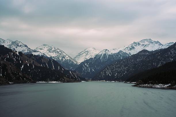 Scenic winter landscape featuring a calm lake surrounded by tall mountains with snowy peaks and dark pine forests under an overcast sky. The image captures the beauty and serenity of untouched nature, ideal for themes of wilderness, travel, tranquility, and natural environment.