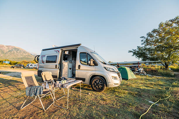 Silver camper van with open door parked on grassy campsite near green tent and motorcycles. Outdoor chairs and table set with cups and cloth. Scenic camping area with mountains and trees.