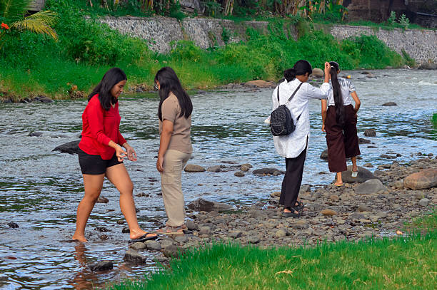 Four teenage girls walk along the shallow river, exploring the rocky stream path while enjoying the wild grasses, fresh greenery, and calm atmosphere of the riverside meadow.