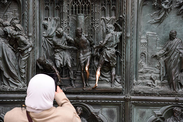The image shows some bronze details at the huge main entrance door. And a women taking a photo with her smartphone.