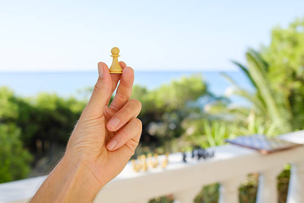 Hand holding a white pawn chess piece on a balcony with a blurred sea view and chessboard in the background, symbolizing strategy, planning, and leisure