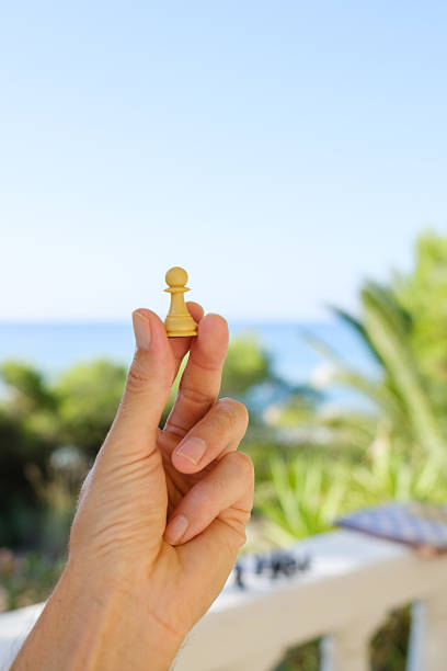 Hand holds a white pawn chess piece against a blurred background of a tropical beach and chessboard, suggesting strategic thinking and leisure