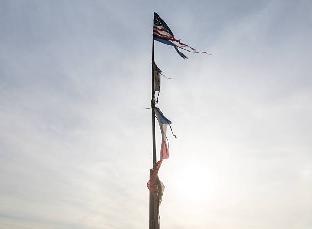 A weathered American flag attached to a pole, with colorful ribbons, set against a bright sky.