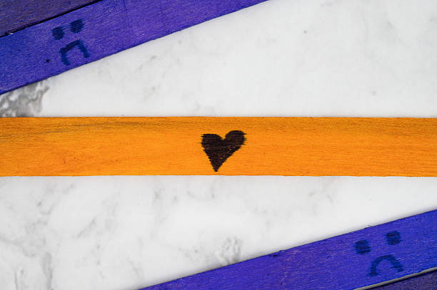 Orange popsicle stick with a black heart drawn on it, placed diagonally across a marble surface, surrounded by purple sticks with sad face icons.