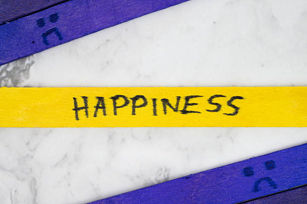 Yellow stick with handwritten word Happiness placed between two purple sticks with sad face drawings on white marble background.