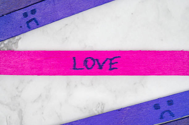 Bright popsicle sticks on a marble surface with the word LOVE written on a pink stick, surrounded by sad face drawings on purple sticks.