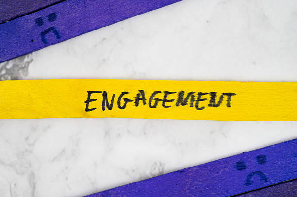 Yellow stick with handwritten word Engagement placed between purple sticks with sad face drawings on white marble surface.