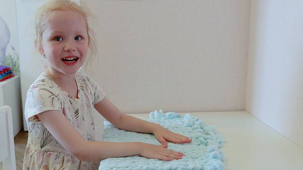 Joyful young girl caressing a plush, light blue blanket on a white table, savoring its softness and cultivating a warm, cozy ambiance