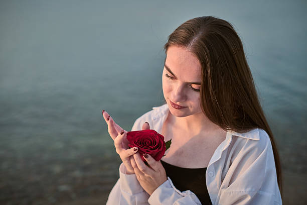Young woman gently holds and gazes at red rose by calm lakeside. Her peaceful expression and serene environment suggest contemplation and tranquility. Soft lighting enhances soothing ambiance