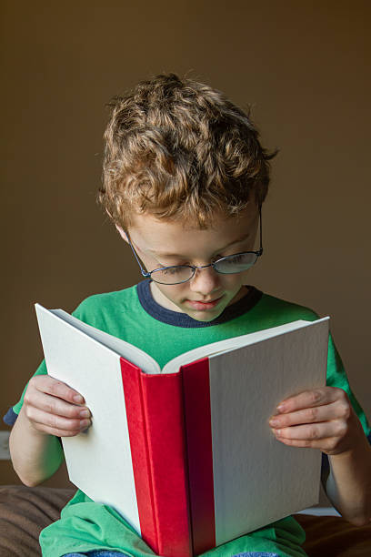 Elementary aged boy reading wearing round glasses and green t-shirt with blue trim sitting on brown cushion with brown wall cropped at waist. Holding blank white book with red spine to add copy.