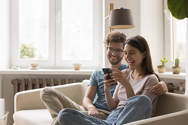 istock Glad young couple hugging on sofa making videocall via cellphone 2234044710