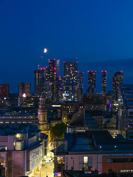A portrait-oriented aerial photograph of downtown Manchester during blue hour. The photograph was created just as the city had begun turning on its lights in readiness for the early evening.