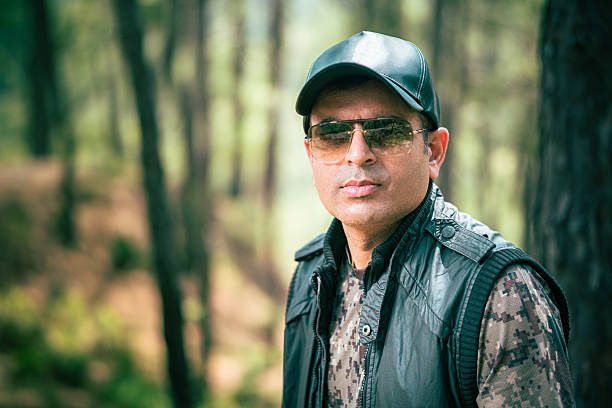 A confident mid-adult Indian man poses for a portrait in a pine forest. He wears sunglasses, a cap, and a vest over a camouflage shirt, standing in tree area while looking at the camera.