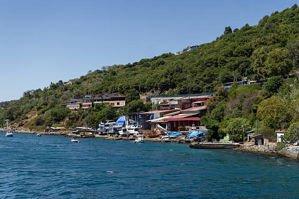 Istanbul, Turkey - September 7, 2025: A boatyard with small fishing boats and repair sheds located on the Bosphorus shoreline, surrounded by green hills and waterfront houses under clear blue skies.