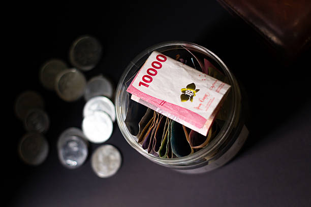 Close-up of a jar filled with Indonesian rupiah banknotes and scattered coins on a dark surface, illustrating savings planning and financial security for future investments.