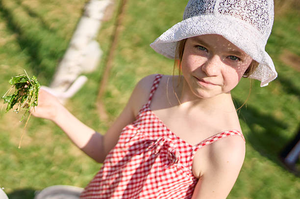 A beautiful elementary age girl dressed in a lace panama and bright summer clothes spends a weekend in a rural cottage. She is cooking a herbal soup in make-believe play. Shooting on a back yard of a country cottage at a sunny spring day