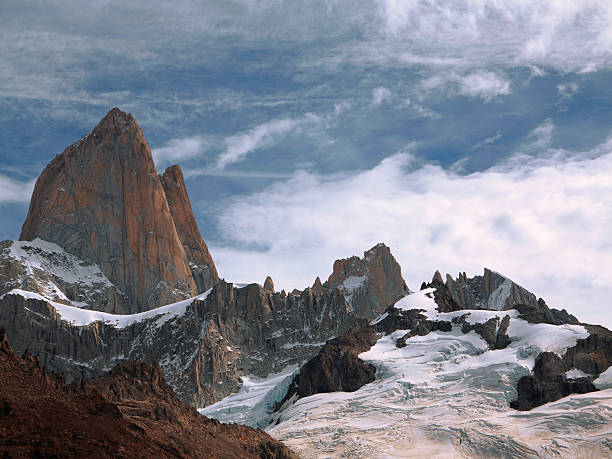 Stunning high-resolution image of a dramatic alpine landscape featuring sharp, snow-covered peaks and a prominent reddish-brown rock spire. The scene captures the raw beauty of glaciated terrain with visible crevasses, steep cliffs, and textured ice formations. Partly cloudy sky adds depth and atmosphere, making this photo ideal for use in travel, adventure, nature, geology, and environmental themes. Perfect for editorial, commercial, or background use in outdoor-related content.