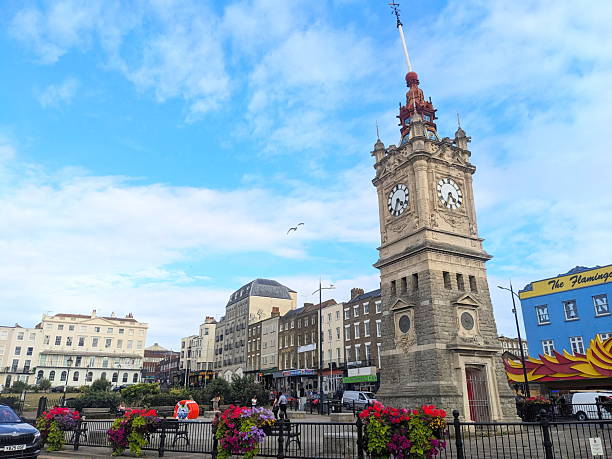 Margate, UK - August 23 2025: The historic stone Jubilee Clock Tower overlooks the vibrant seafront promenade, with colourful flowers, traditional buildings, and a bright blue summer sky