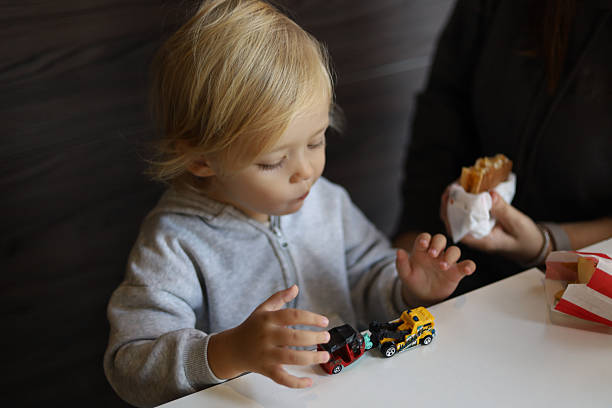 Three-year-old boy playing with toy cars while mother feeds him a burger