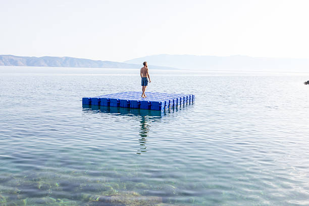 Individual lying on a blue floating platform in clear calm sea water near rocky shore with pine trees. Peaceful coastal scene with sunlight reflecting on water. Relaxation and solitude by the sea.
