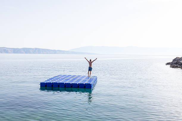 Individual lying on a blue floating platform in clear calm sea water near rocky shore with pine trees. Peaceful coastal scene with sunlight reflecting on water. Relaxation and solitude by the sea.