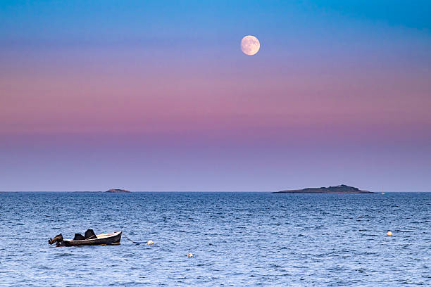A small boat gently floats on the ocean under a vibrant twilight sky. The Corn Moon shines brightly, casting a serene glow over the water. Two small islands appear in the distance, adding depth to the tranquil scene in Beverly, Massachusetts.