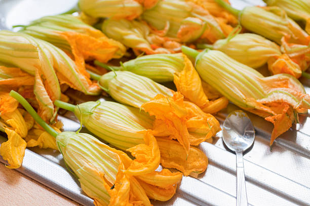 Vibrant yellow edible zucchini blossoms on a metallic tray with a spoon, showcasing fresh green stems and delicate petals. Perfect for culinary use, this close-up highlights their natural beauty and texture in a kitchen setting