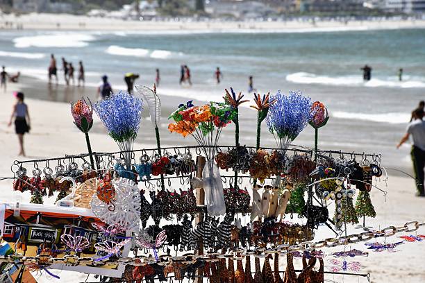 Landscape 16:9 handmade bead crafts and souvenirs on beach stand with ocean and people in background.