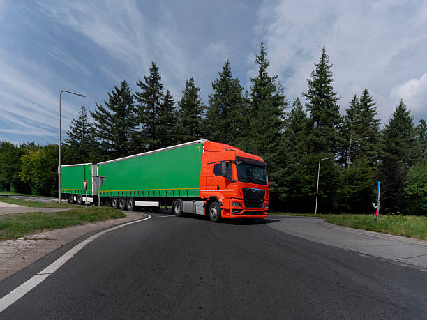 Orange truck with green trailer navigating road through evergreen forest with cloudy sky