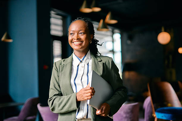 Smiling confidently while walking through a modern office, a young African American businesswoman holds a laptop, embodying optimism and professionalism in her career journey