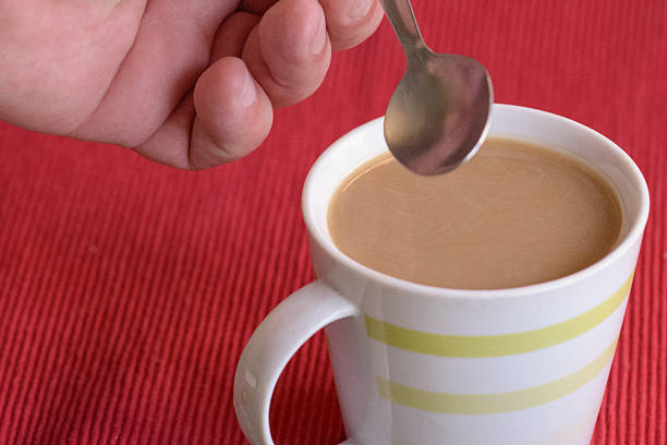 A hand stirs a cup of coffee with a spoon on a vibrant red tablecloth during the morning.