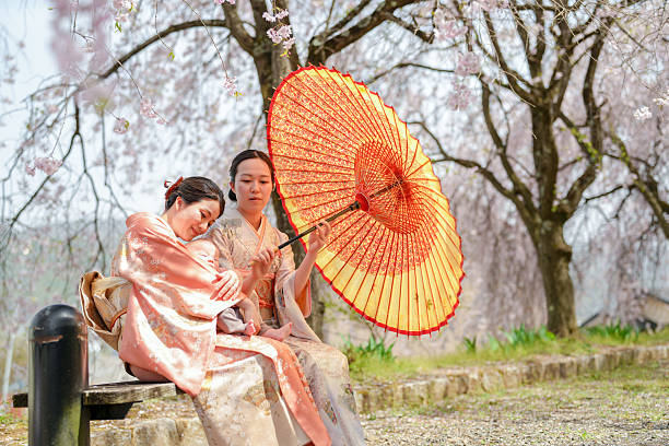 Two women are sitting on a bench under a red and yellow umbrella. They are dressed in traditional Japanese clothing, and the scene takes place in a park with cherry blossoms in the background