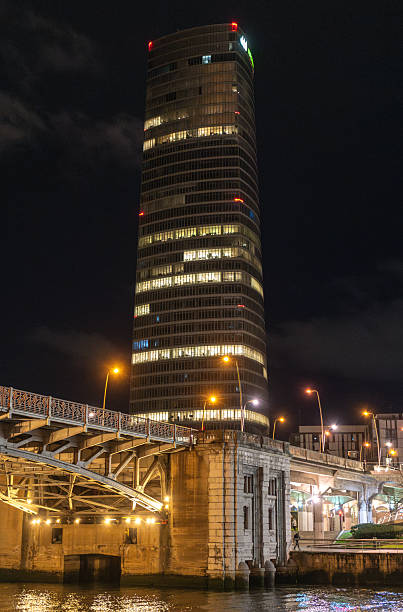 Bilbao, Spain, March 28, 2024: Iberdrola Tower in Bilbao at night behind illuminated bridge with stone support structure and street lamps, editorial image