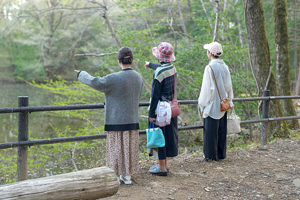 Three women are standing on a path near a pond, looking at something in the distance. They are wearing hats and carrying handbags