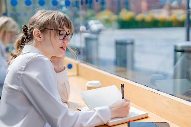 istock Young woman writing in notebook while sitting by window in a bright cafe during daytime 2234242950
