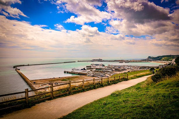 Scenic view over the busy harbour of Dover in Kent, England, with ferries docked at the terminal and protective sea walls stretching into the English Channel. The photo, taken from the cliffs above, highlights the port’s strategic position as the main gateway between the United Kingdom and continental Europe. The dramatic sky, calm sea, and iconic White Cliffs in the background complete the coastal landscape.