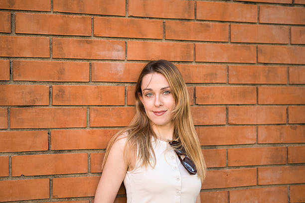Portrait of a young woman standing against a brick wall, smiling while looking at the camera
