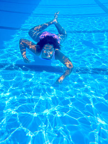 Young girl swimming underwater in a clear blue swimming pool, wearing goggles and a swimsuit, with sunlight reflections dancing across her skin and the water. Captured in a vibrant summer setting, the image conveys fun, motion, and the joy of aquatic activities.