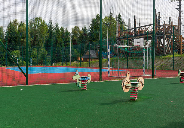 Children’s playground with spring riders in front of a fenced sports field and basketball goal. Surrounded by greenery, the area combines play and exercise, offering fun and activity in a safe environment.