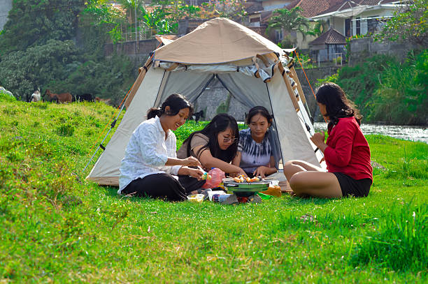 A girl spreads butter on skewers before grilling, while others inside the tent prepare more sausages and bowls for the outdoor riverside barbecue.