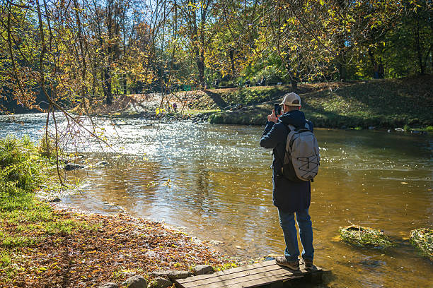 istock Tourist man with a backpack taking a picture with his smartphone by a river in a park during a sunny autumn day 2234273918