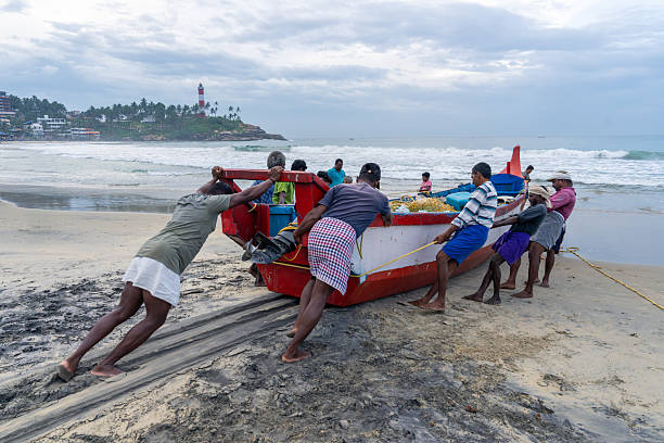Local fishermen work together to pull a red wooden fishing boat ashore on the beach in Kovalam, Kerala, India. The men are barefoot and using thick ropes and physical strength to drag the vessel over the wet sand, their movements perfectly synchronized. The sky is overcast after the early morning catch, and in the background, the iconic red-and-white lighthouse of Kovalam stands on a lush green hill surrounded by palm trees. The scene captures the rhythm of coastal life, teamwork, and the daily labor of small-scale fishing communities along the Arabian Sea. This documentary-style image highlights traditional fishing practices, physical effort, and the human connection to the sea.