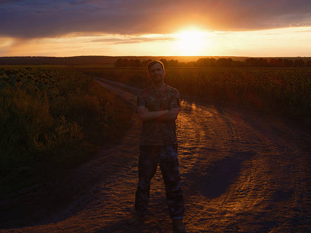 Soldier in camouflage uniform stands on dirt road in sunflower field at sunset. The peaceful rural landscape is contrasted with the military presence, conveying the realities of war. War in Ukraine.