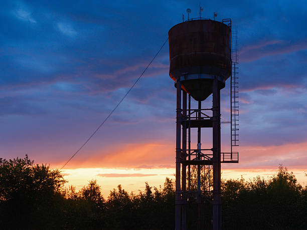 Rusty old water tower stands in silhouette against a dramatic sunset sky. The vibrant colors of the clouds create a beautiful, tranquil rural scene at dusk.