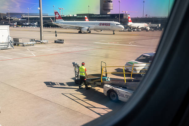 Zurich Airport, Switzerland, August 25, 2025. Luggage handler loading a suitcase on the automated belt into the plane. Airplane window seat view, sunny day